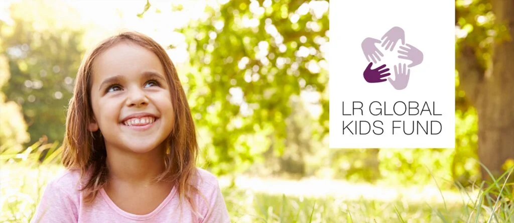 Petite fille souriante assise dans l’herbe, à côté du logo LR Global Kids Fund.