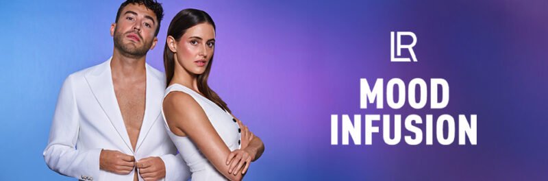 Un homme et une femme posent pour une photo promotionnelle du nouveau parfum "B Mood Infusion" de LR Health and Beauty. L'homme est à gauche, portant une chemise blanche ouverte, et la femme est à droite, portant un haut court et une jupe marron. Le nom de la marque, en lettres violettes, est au centre.